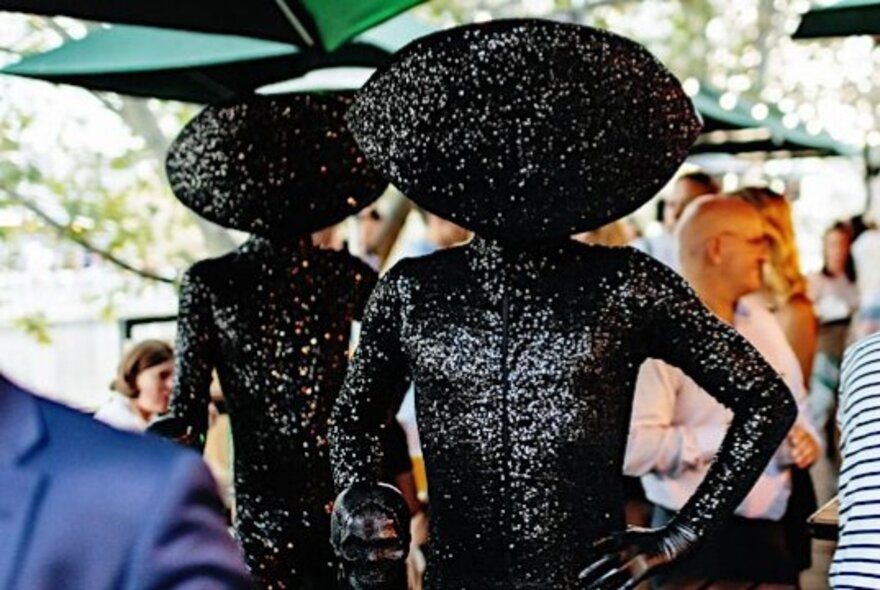 Figures wearing shiny black catsuits with face and head coverings, standing with hands on hips in an outdoor beer garden setting.