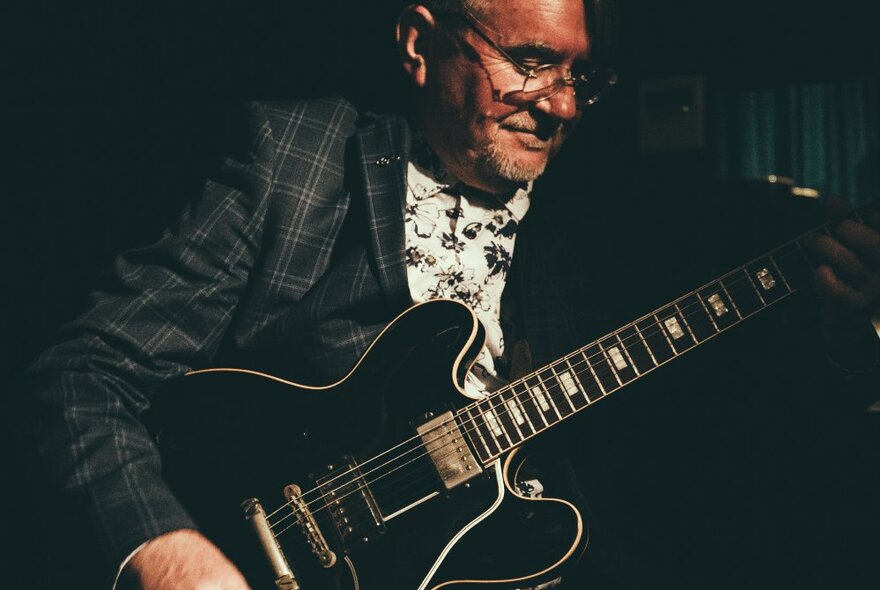 Musician Mark Morand with his black Gibson electric guitar, wearing a suit jacket and white shirt.