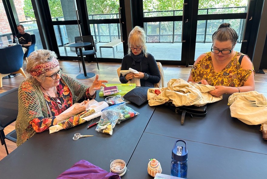 A group of people seated around a large table beside large windows with sewing projects and materials. 