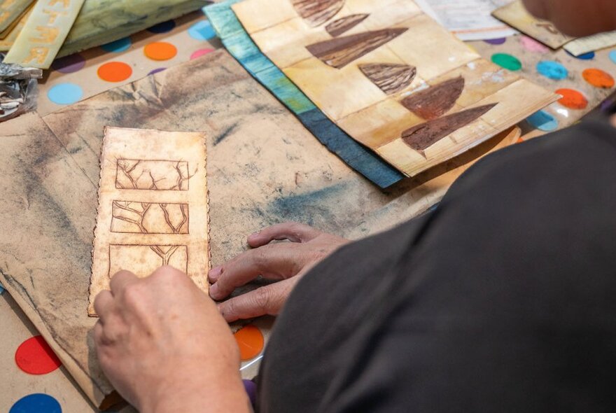 A person's hands working on a piece of brown paper with etched designs, surrounded by other similar craft materials.