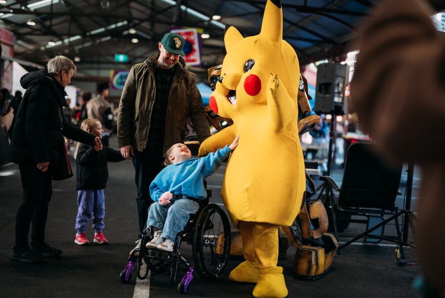A person wearing a life size yellow Pikachu character costume interacting with a young child in a pram, while an adult looks on, at the Queen Victoria Market.