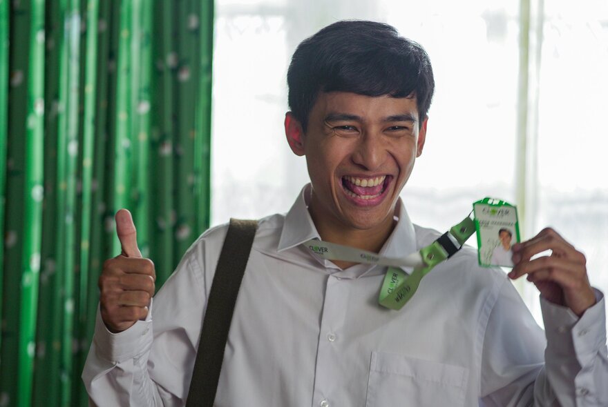 A young man with dark hair and a wide smile gives a thumbs-up while holding up an identification lanyard; a still from a movie.