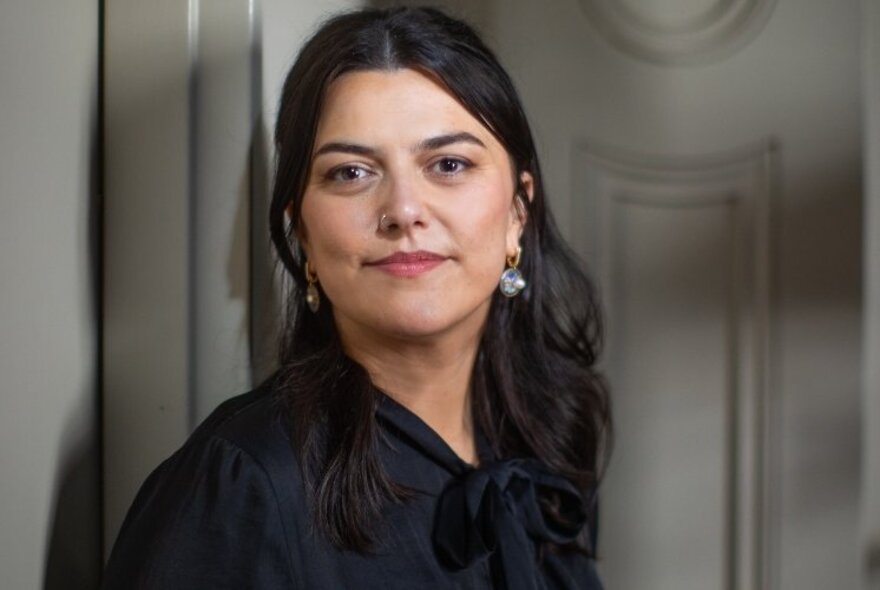 A woman with straight dark hair, sliver earrings, and wearing a black silk blouse, standing in a room with white walls.