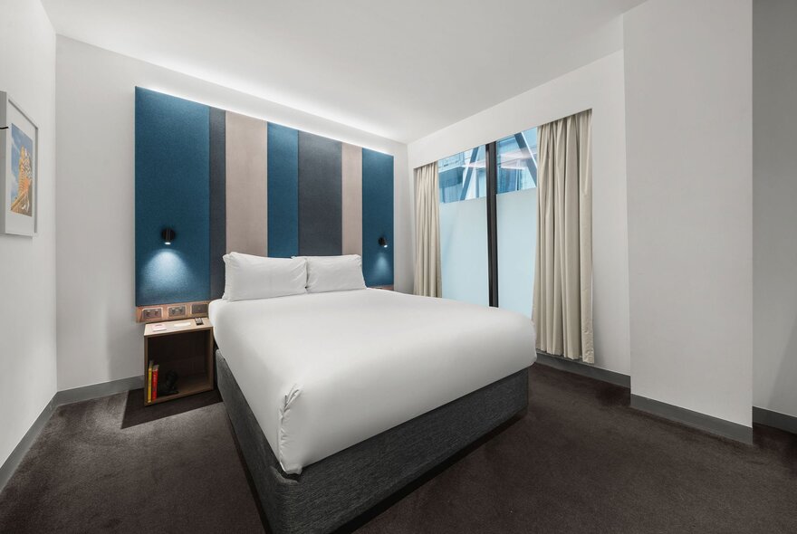 A contemporary hotel room at the ibis Styles Melbourne Southbank featuring a queen-sized bed with a white duvet and blue feature panelling.