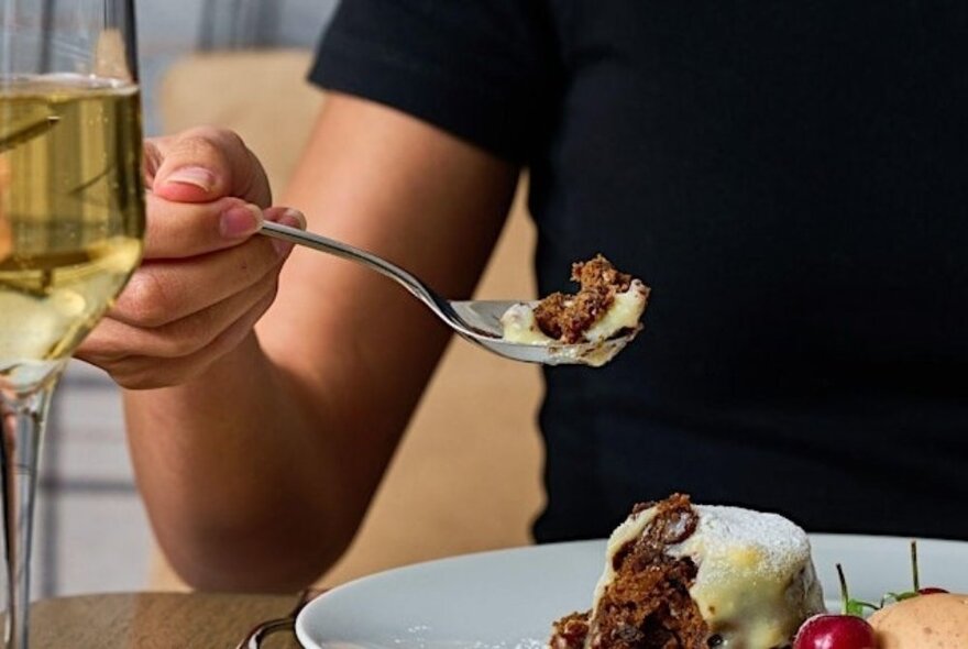 Person moving a spoonful of Christmas pudding from a dish towards their mouth, glass of wine to the side. 