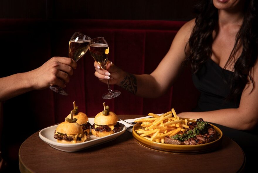 Two people clink glasses over a plate of sliders and another of chips. 