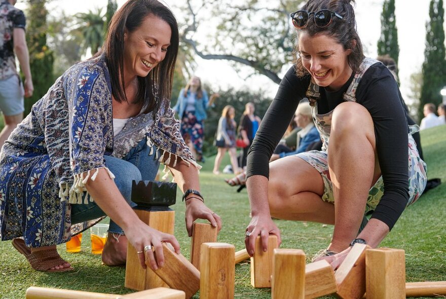 Two smiling friends setting up a lawn game with multiple wooden blocks, other people walking around on the lawns in the background.