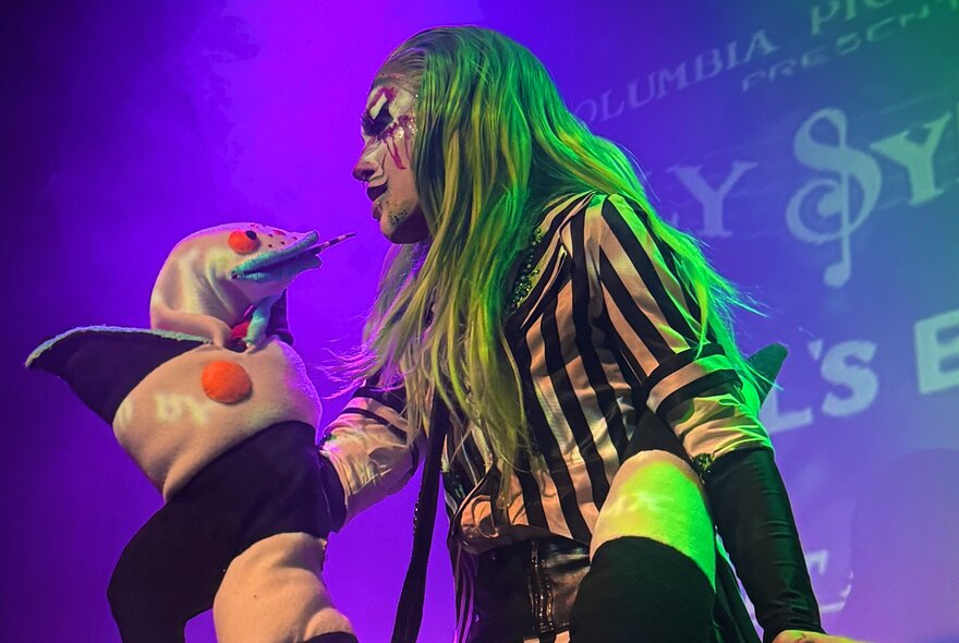A drag performer on stage with a hand-held puppet in their arms.