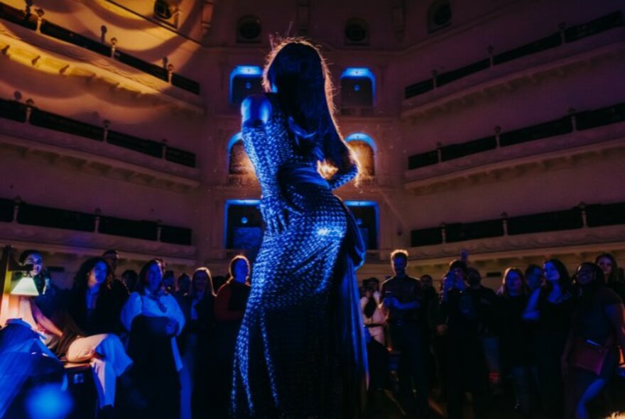 A drag queen dressed in dark blue sequins, in front of an audience at the State Library Victoria.