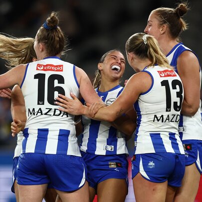 North Melbourne AFLW players embrace in celebration on the football field.