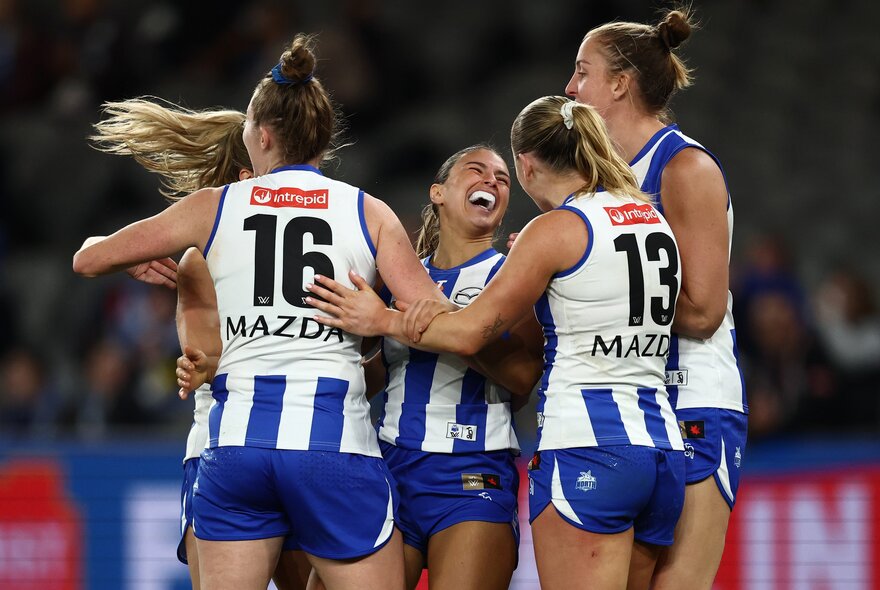 North Melbourne AFLW players embrace in celebration on the football field.