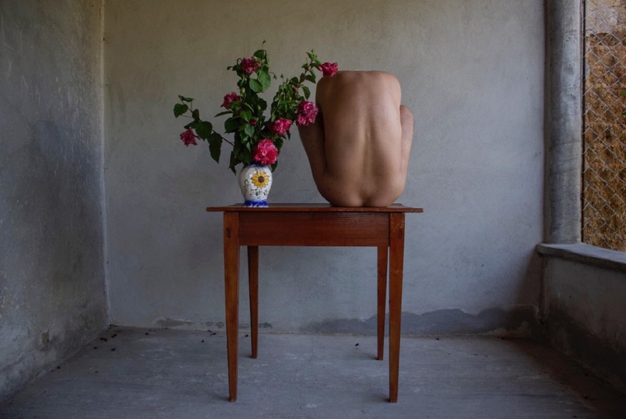 Self-portrait of a nude artist huddled on top of a wooden table, next to a vase of pink flowers in an otherwise empty concrete room 