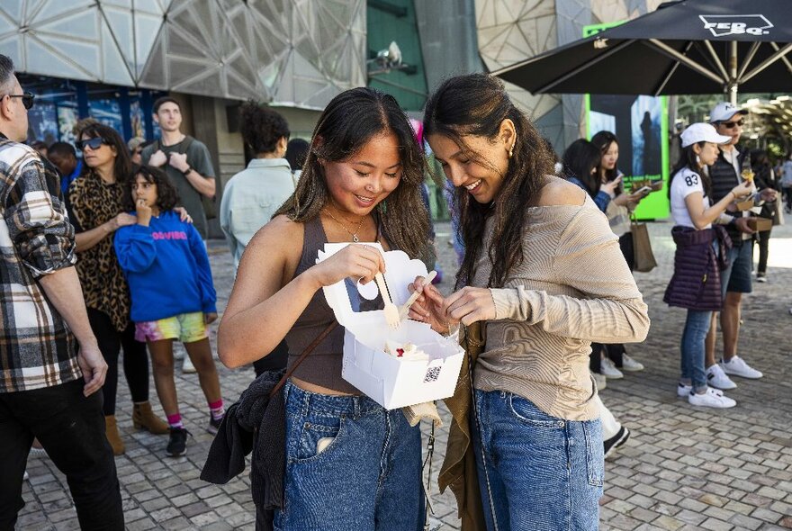 Two people in Fed Square holding a box of cakes, surrounded by people.