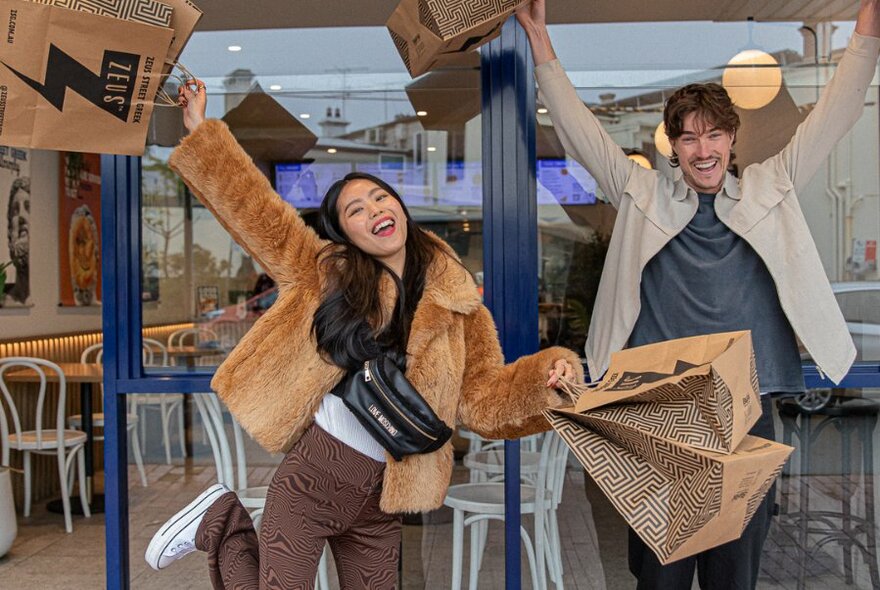 Two people holding paper bags out front of shop front, cheering.