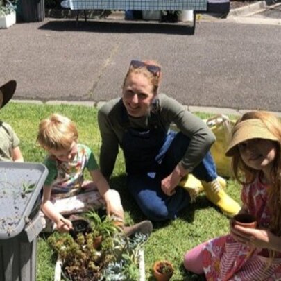 An adult and three young children potting or tending to small plants in a sunny, grassy area at the Royal Botanic Gardens Melbourne.