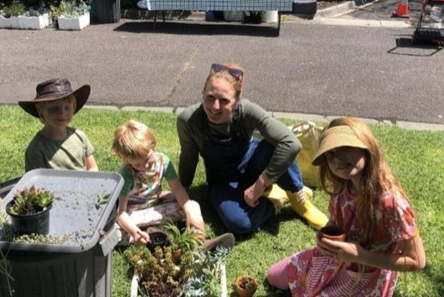 An adult and three young children potting or tending to small plants in a sunny, grassy area at the Royal Botanic Gardens Melbourne.