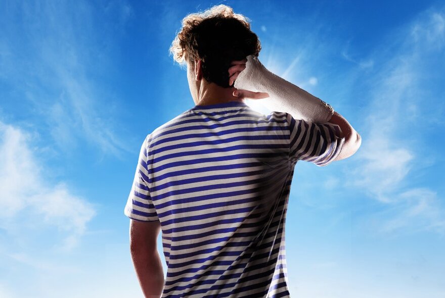 The back of a young adult who has their right arm in a plaster cast and positioned behind their head, wearing a blue and white striped t-shirt, and standing against a blue sky background.