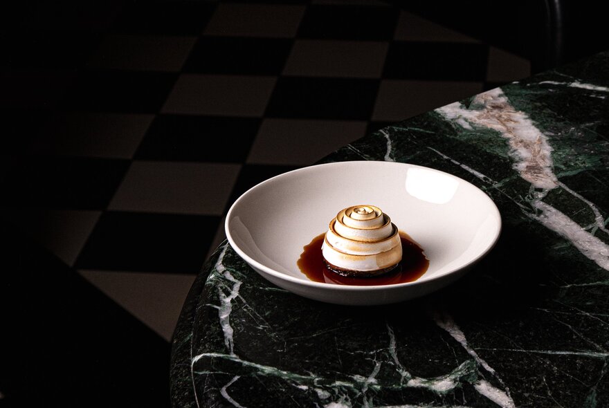 A dessert wth spiralled torched meringue in a dark sauce in a white bowl on a deep green marble table. 