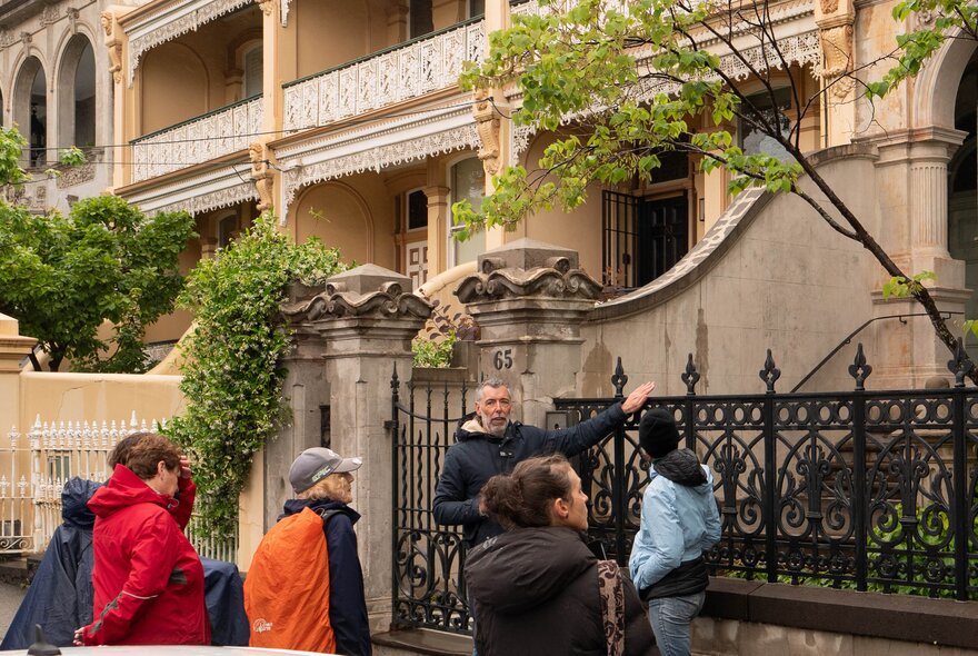 A tour guide leading a walking street tour, pointing to a terrace house, with a few people standing around listening.
