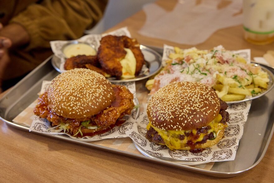A tray with two burgers in sesame seed buns with chicken poking out and sauce oozing down, loaded fries and patties. 