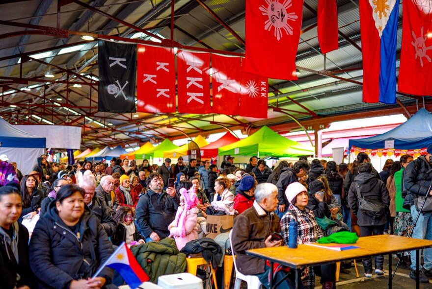 A crowd of people sitting and standing at the Queen Victoria Market, with Filipino flags above them,