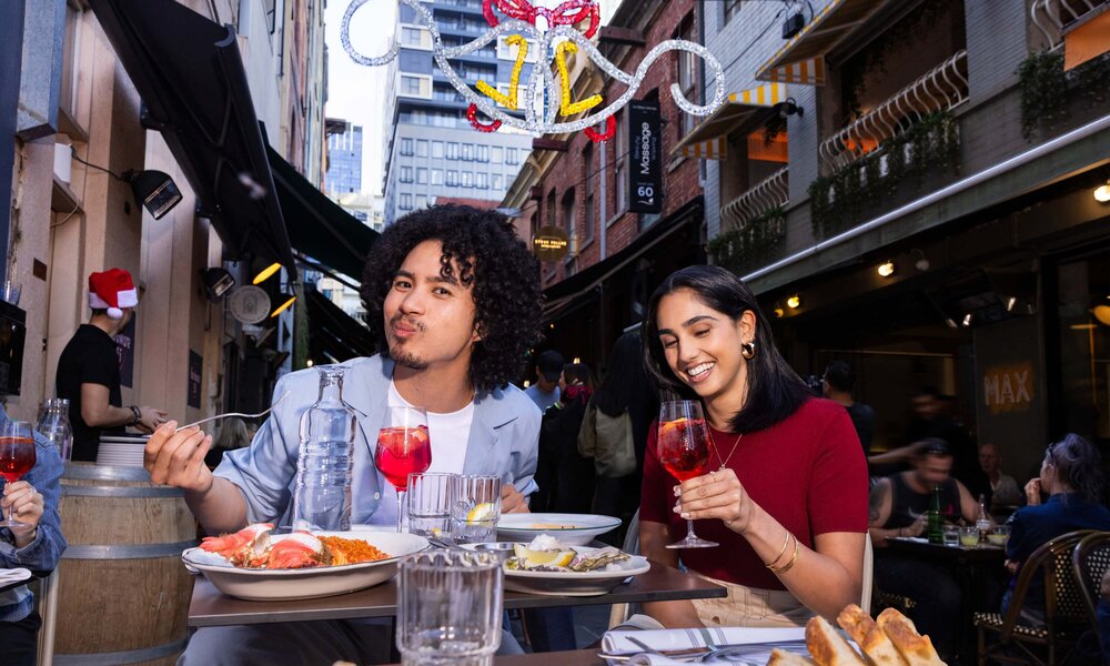 A man and woman eating outside in a laneway under Christmas decorations. 