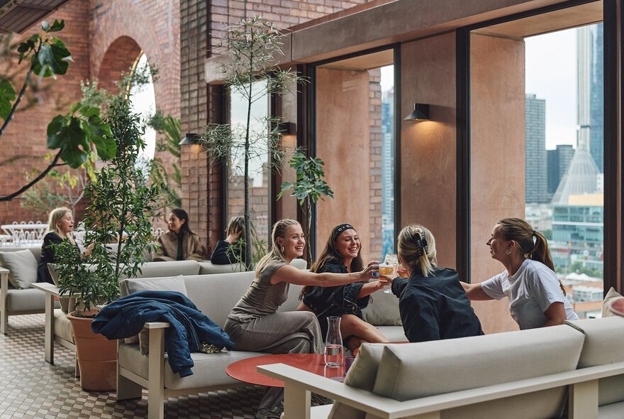 Four women clink glasses in a toast while sitting on sofas in a rooftop bar during the day. 