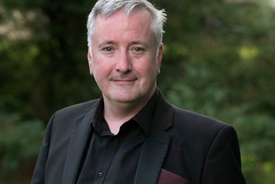 Arts journalist and broadcaster Richard Watts, a middle-aged man wearing a dark suit, posed outside with greenery behind him.