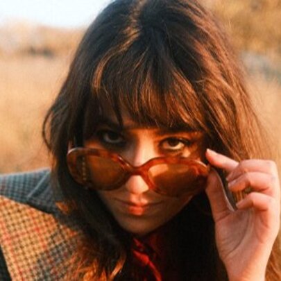 Singer Gabriella Cilmi at sunset in a field, pulling her sunglasses down to the end of her nose. 