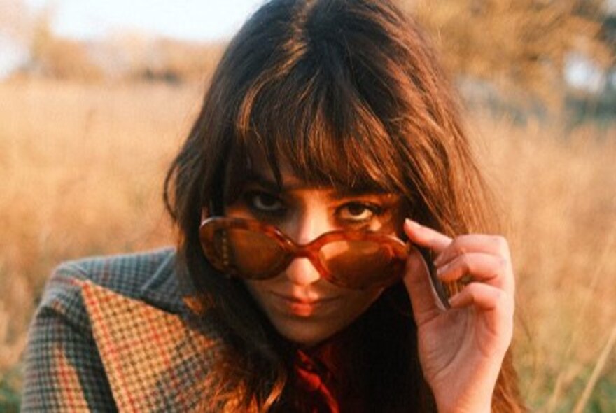 Singer Gabriella Cilmi at sunset in a field, pulling her sunglasses down to the end of her nose. 