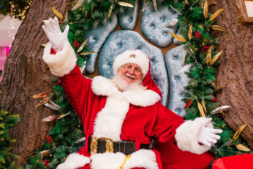 A happy and smiling Santa with his arms outstretched sitting in a decorated Christmas tree chair.