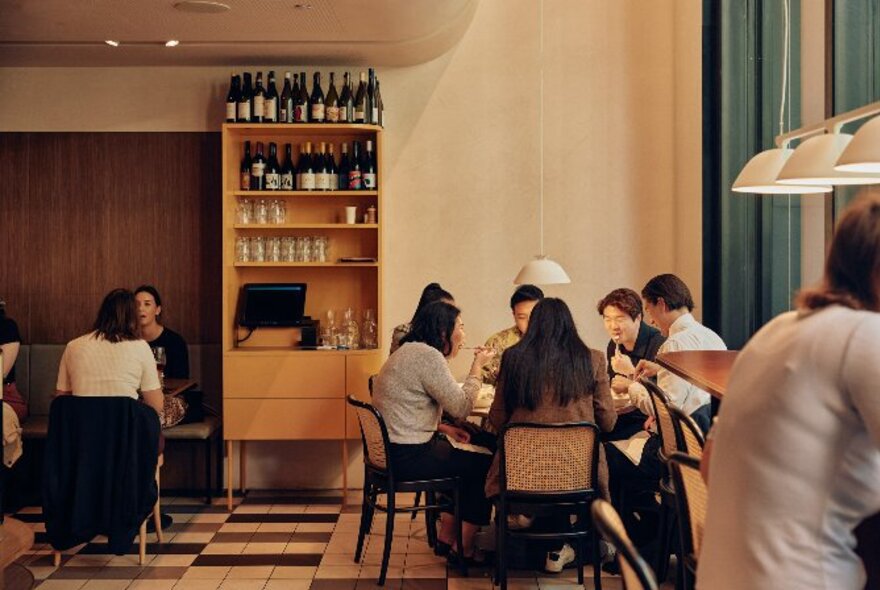 Diners seated in a restuarant space wiht a small shelf of wine. 