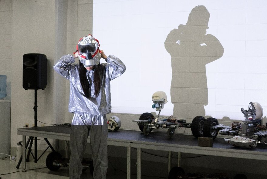 A woman with long hair in a silver track-suit putting on a helmet in front of a white wall with her shadow projected on it.