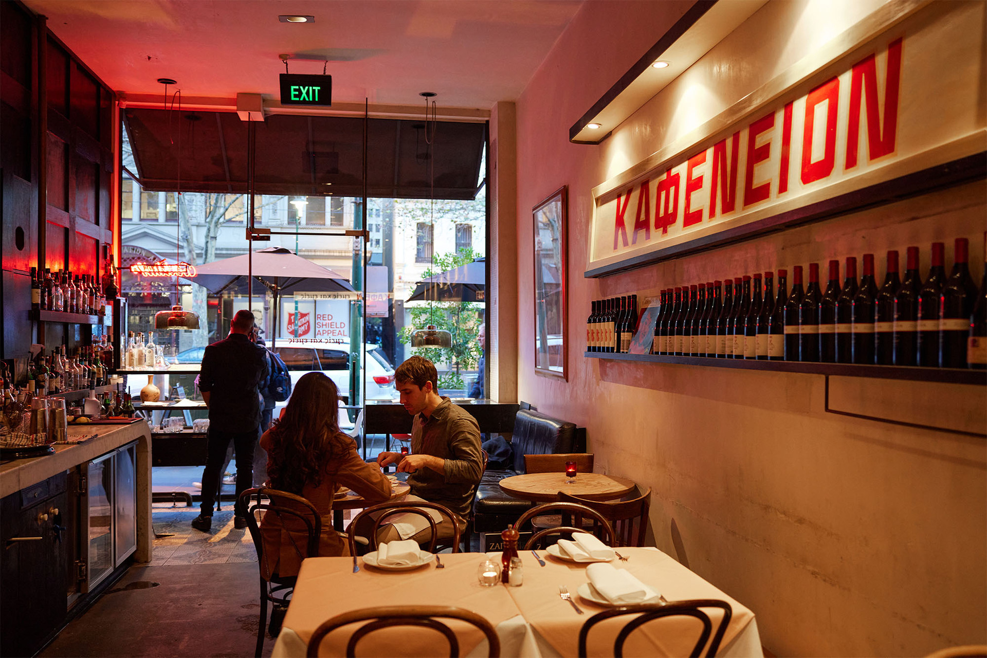 Two people at a Greek restaurant enjoying a meal. There is a large red painted sign on the wall saying Kafeneion.