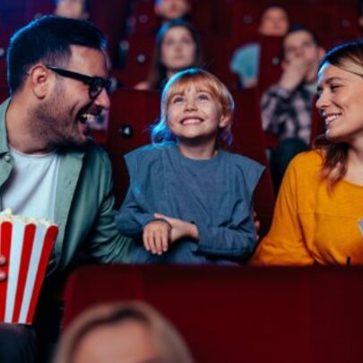 Parents and child seated in cinema seats, parents holding large striped containers of popcorn, the child in the middle smiling up at the screen.