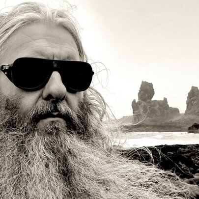 Headshot of sound and light artist Robin Fox, a man with a thick long white beard and long hair, wearing sunglasses, standing in front of a rocky coastline and water.