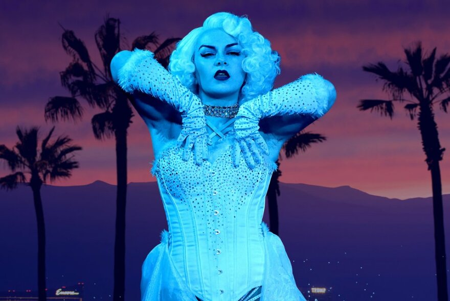 Cabaret performer wearing white gloves and bustier against a sunset landscape with palm trees.