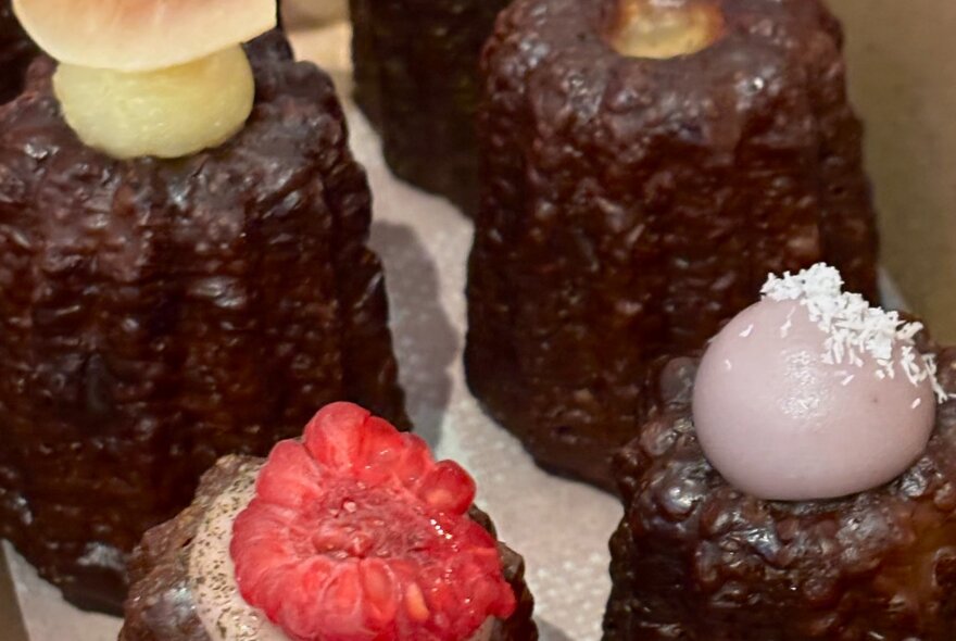 Four chocolate canelé, pudding-like mini desserts, with sliced fruit or dollops of icing on top.