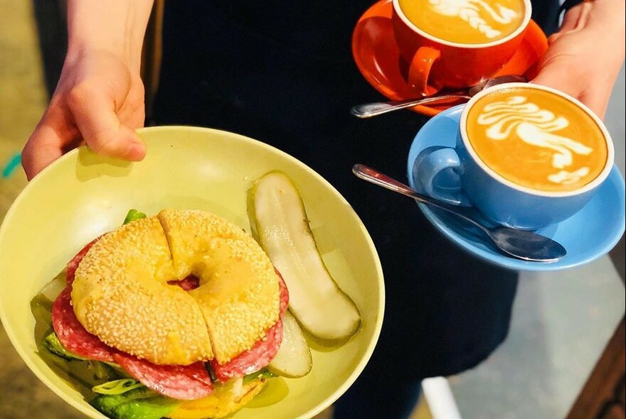 Waiter holding two cups of coffee and a yellow bowl with a pastrami bagel with pickle.