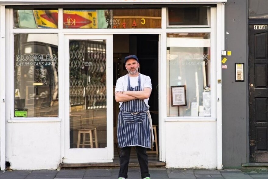 Chef standing with arms folded, wearing striped navy apron, in front of a streetside restaurant.