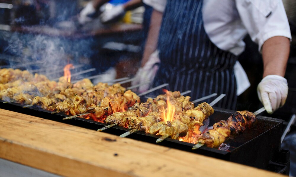 A chef cooking meat skewers over a flaming grill. 