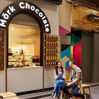 Two friends are seating outside a small cafe kiosk there is a sign saying mork chocolate