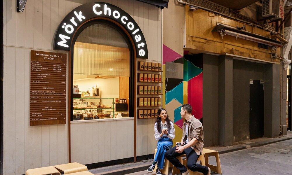 Two friends are seating outside a small cafe kiosk there is a sign saying mork chocolate