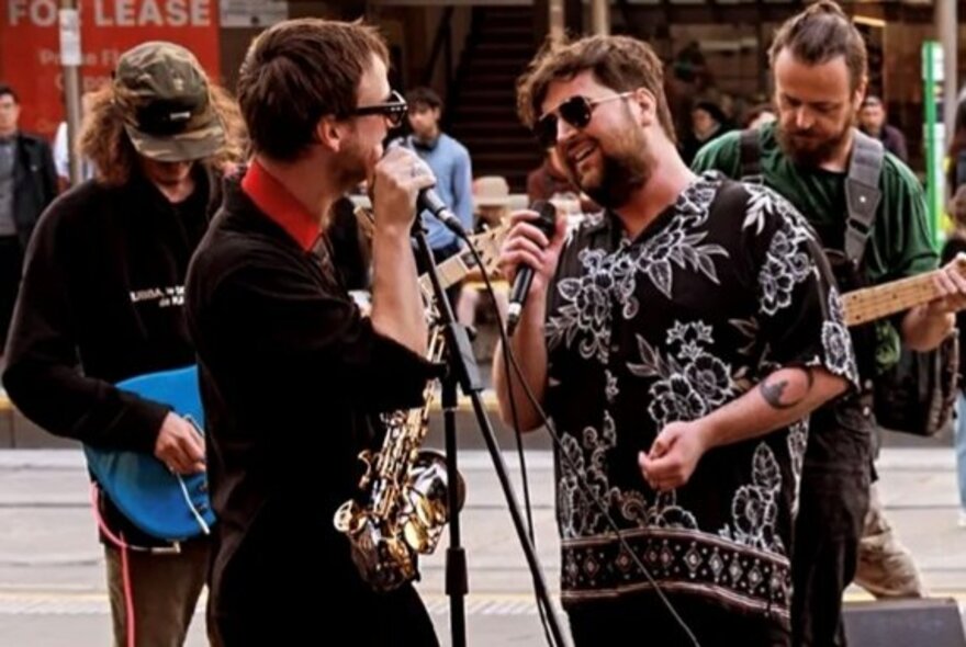 Two street performers singing into microphones, with two guitarists playing music behind them.
