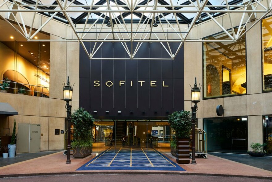 The exterior undercover foyer at Sofitel Melbourne on Collins, with two ornamental lamp-posts and plants lining each side of a blue carpet leading to glass entrance doors, with the words SOFITEL, written in large letters above the doors.