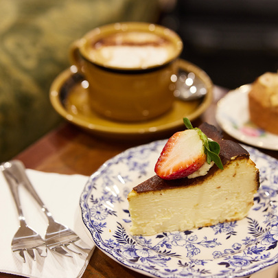 A slice of cheesecake on a decorative blue plate, behind it is a cup of coffee and a friand 