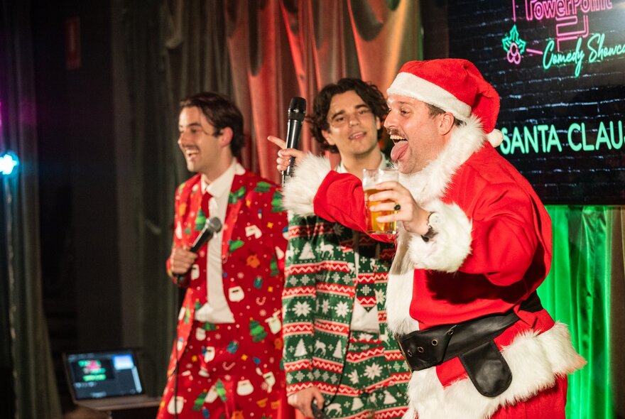 Two people on stage, wearing festive-themed outfits, plus Santa holding a pot of beer and with his tongue poking out, performing in a small theatre room.