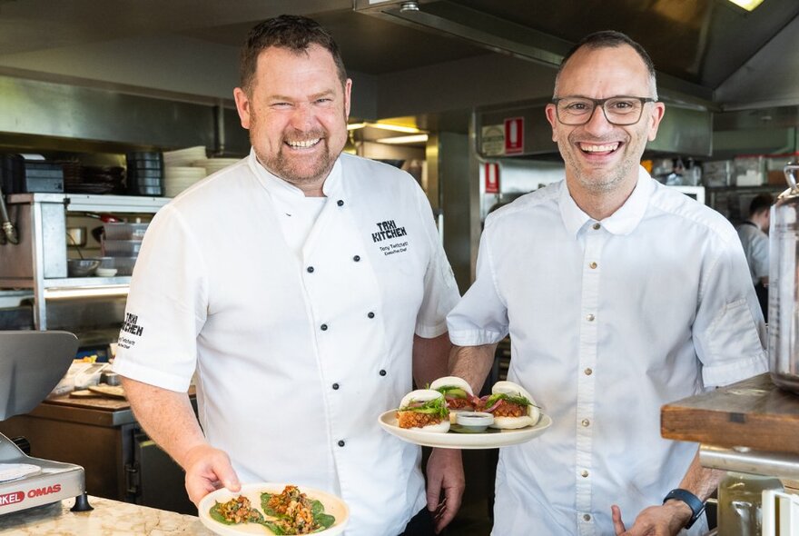 Two chefs wearing their white chef's uniforms, smiling and holding up plates of food, in a commercial restaurant kitchen.