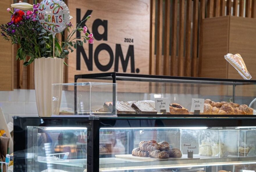 A shop counter with pastries and a vase of flowers with a celebratory balloon.