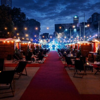 An outdoor alpine village pop-up event at Fed Square at night, featuring private wooden chalets, string lights, and a red carpet.